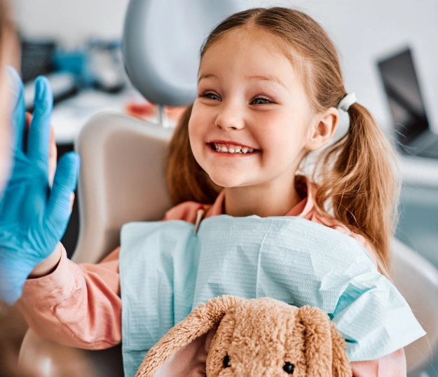 Young girl smiling in dental chair holding teddy bear with dentist in blue gloves.