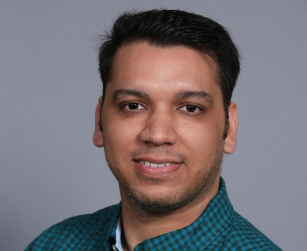 Professional headshot of male dentistry team member wearing teal checkered shirt against gray background.