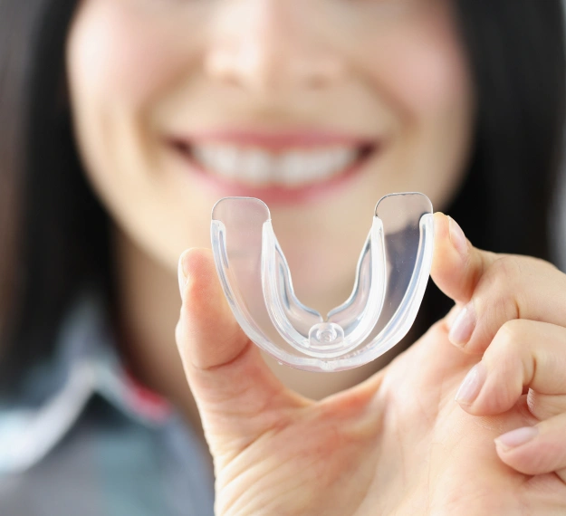 Woman smiling while holding clear orthodontic retainer or aligner tray in her hand.