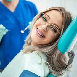 Smiling patient wearing glasses in dental chair with dentist in blue scrubs nearby.