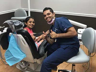 Pediatric dentist in blue scrubs demonstrates proper tooth brushing technique to young patient in dental chair.