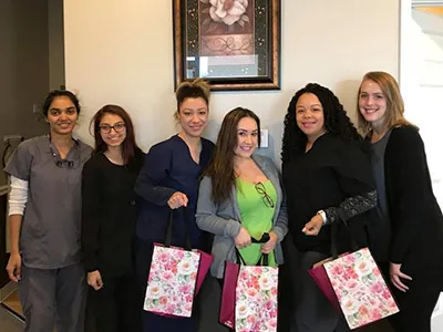 Six women standing together indoors holding pink gift bags, smiling at camera in dental office setting.