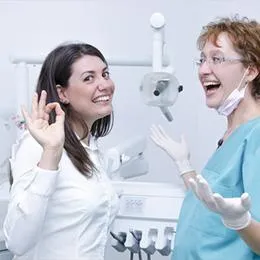Two female dental professionals smiling and communicating in a modern dental office operatory.
