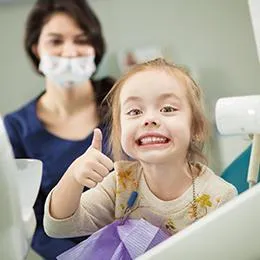 Young girl giving thumbs up at dental office with masked dentist in background.