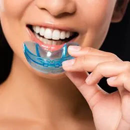Woman inserting blue custom teeth whitening tray into mouth against black background.