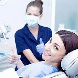 Smiling female patient in dental chair with masked dentist holding tools in modern clinic setting.